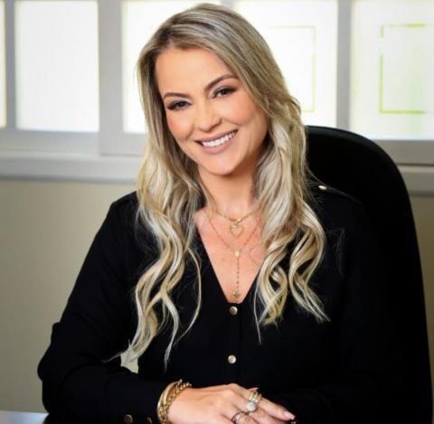 Mulher adulta de pele clara, sentada em uma cadeira, em ambiente interno bem iluminado. Ela tem cabelos longos, loiros, levemente ondulados, usados soltos, e sorri de forma acolhedora para a câmera. Veste uma blusa preta de mangas longas, com botões frontais, e usa colares delicados, pulseiras e anéis dourados. As mãos estão apoiadas uma sobre a outra à frente do corpo. Ao fundo, há uma parede clara com janelas, transmitindo um ambiente profissional e organizado.